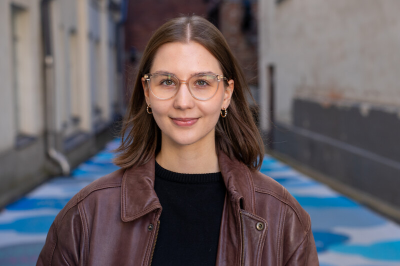 Ruskeahiuksinen, silmälasipäinen nuori nainen katsoo hymyillen kameraan. Taustalla siniseksi maalattu katukivetys.