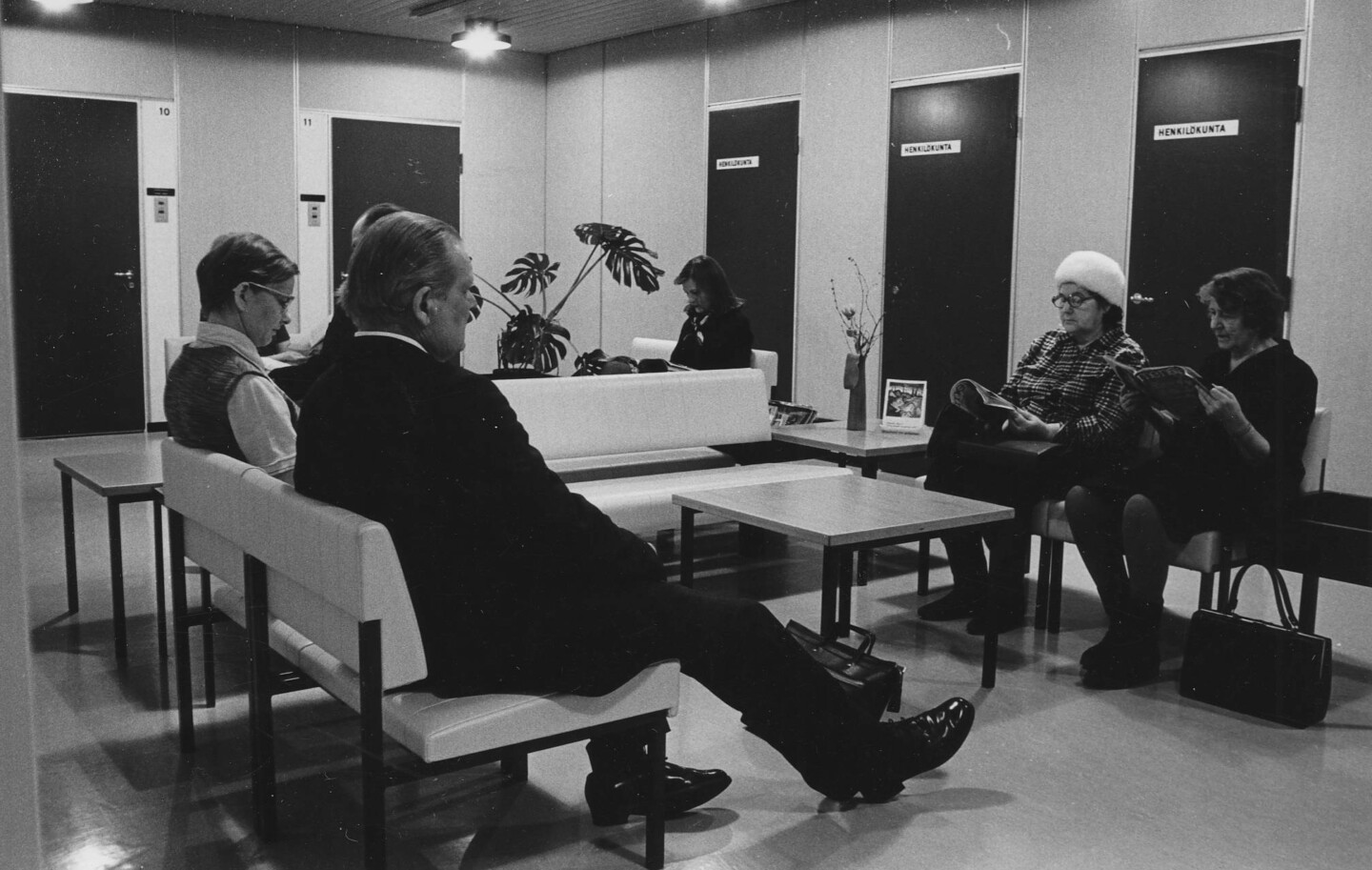 Mustavalkoinen kuva 1970-luvun odotustilasta, jossa viisi ihmistä istuu penkeillä ja odottaa vuoroaan.
