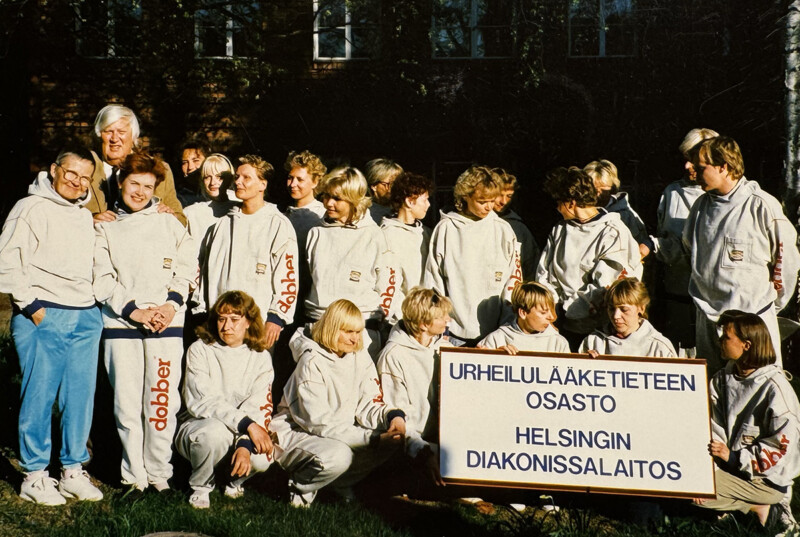 Ryhmä ihmisiä ulkona, kaikki pukeutuneina vaaleisiin urheiluasuihin, osa seisoo ja osa istuu nurmikolla, eturivissä kaksi henkilöä pitelee kylttiä, jossa lukee "Urheilulääketieteen osasto Helsingin Diakonissalaitos".