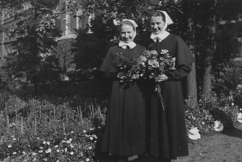 Kaksi hymyilevää diakonissaa mustavalkoisessa valokuvassa, jotka seisovat ulkona puutarhassa ja pitävät kukkakimppuja käsissään.