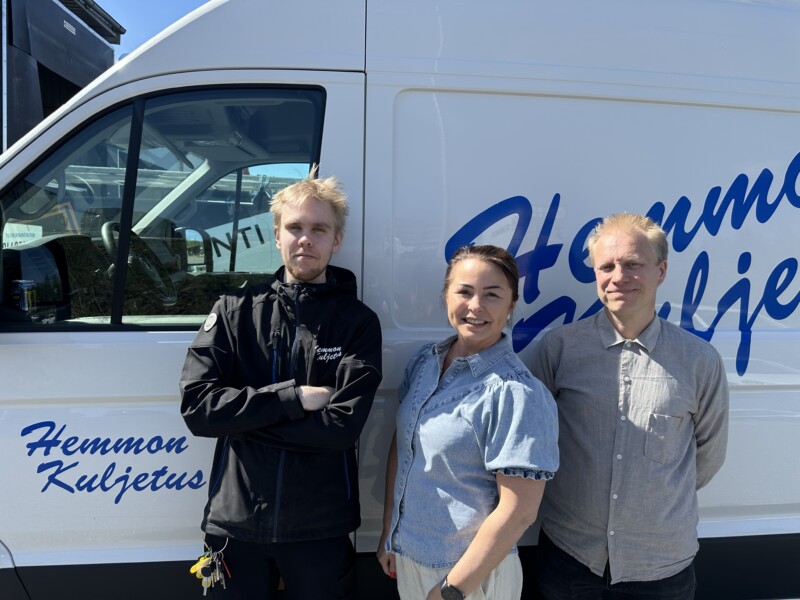 Kolme ihmistä seisoo valkoisen pakettiauton edessä ja katsoo kameraan hymyillen.