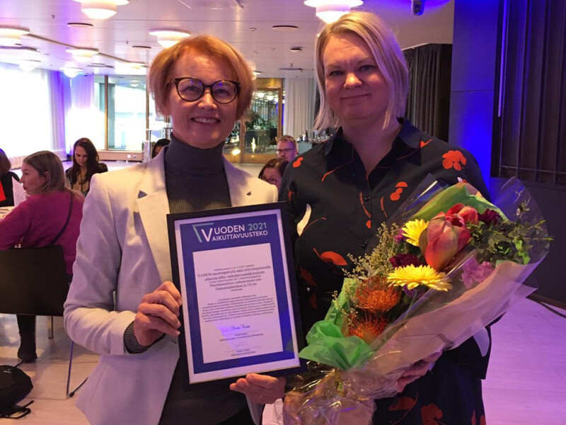 Two women standing indoors, one holding a framed certificate and the other holding a bouquet of flowers, both smiling at the camera.