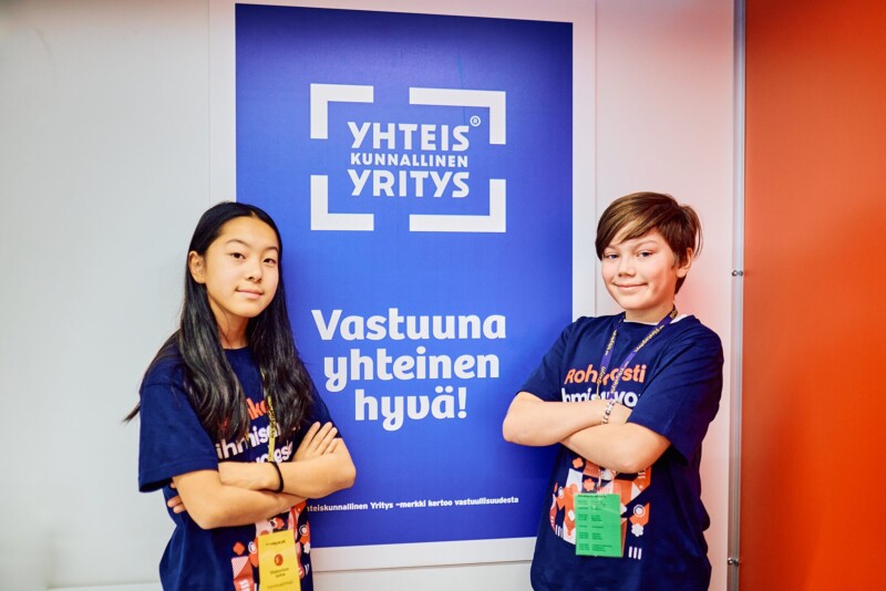 Kaksi alakouluikäistä lasta seisoo hymyillen katsoen kameraan. Heillä on yllään Diakonissalaitoksen t-paidat ja heidän takanaan on juliste, jossa lukee: Yhteiskunnallinen yritys: vastuuna yhteinen hyvä!".