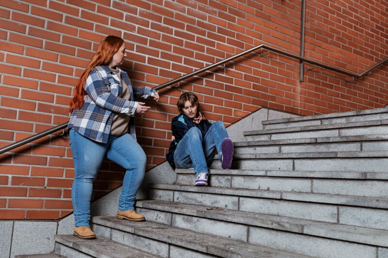 Nuori ja aikuinen keskustelevat. Nuori istuu betoniportailla, aikuinen seisoo hänen vieressään.