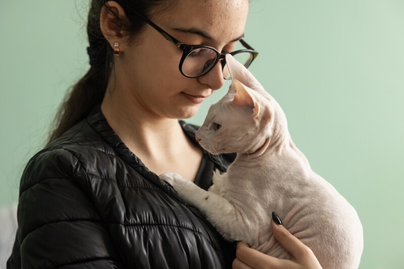 A young woman wearing glasses and a black jacket gently holding and looking at a white Sphynx cat against a plain light green background.