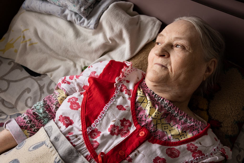 Värikkääseen vaatteeseen pukeutunut vanhempi nainen makaa sängyssä katsoen kohti ikkunasta tulevaa valoa.