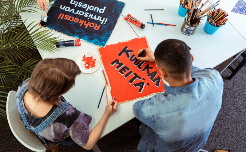Two people sitting at a table engaged in painting colorful signs with Finnish text, surrounded by art supplies like paintbrushes, pencils, and markers.
