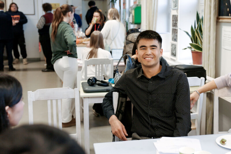 Mustaan huppariin pukeutunut mies istuu pöydän ääressä ja katsoo hymyillen kameraan.