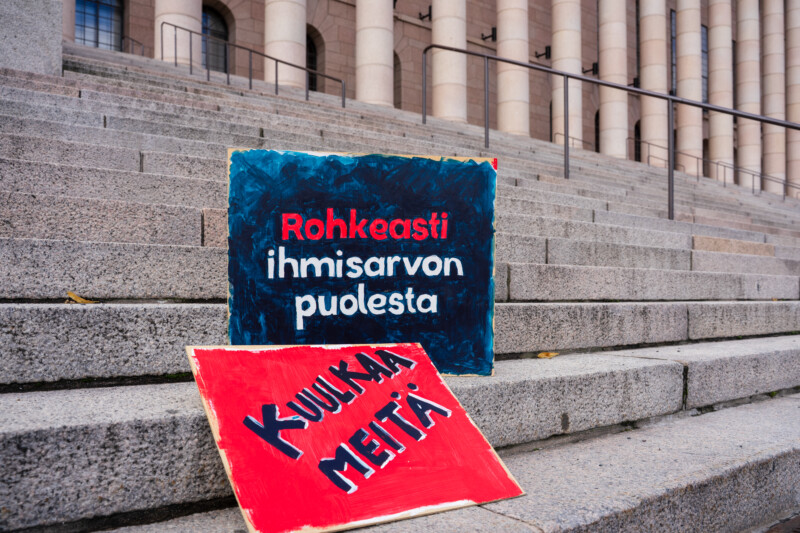 Eduskuntatalon portailla kaksi kylttiä, joissa lukee "Rohkeasti ihmisarvon puolesta" ja "Kuulkee meitä!".