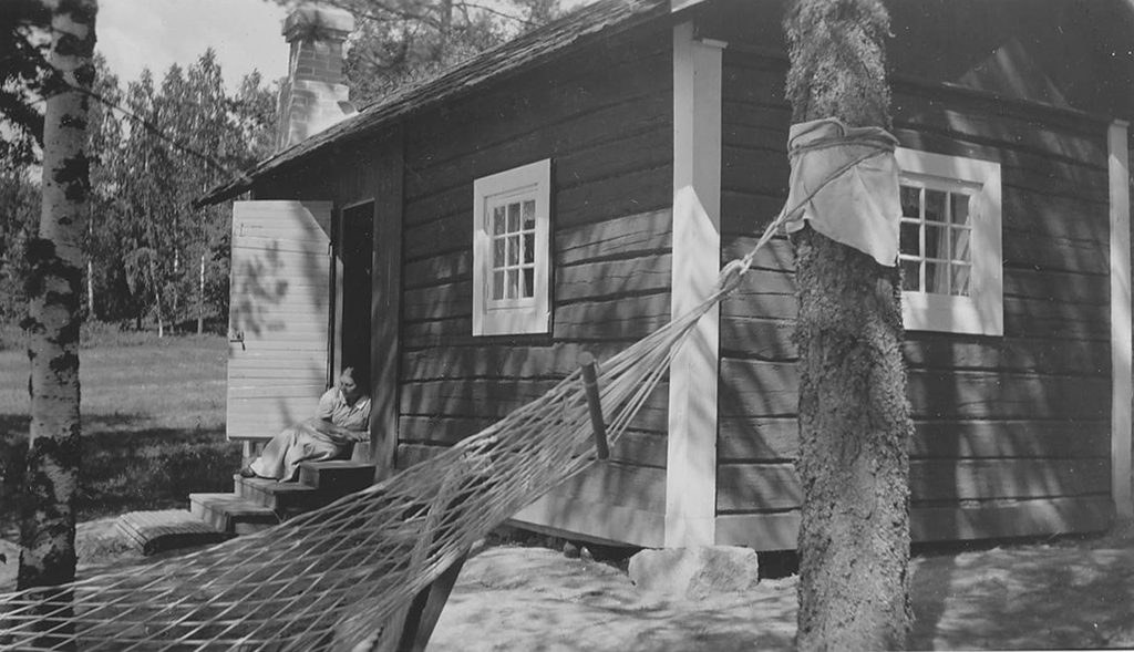 Mustavalkoinen kuva, jossa mökki ja edessä roikkuva riippumatto, taustalla istuu henkilö mökin portailla.