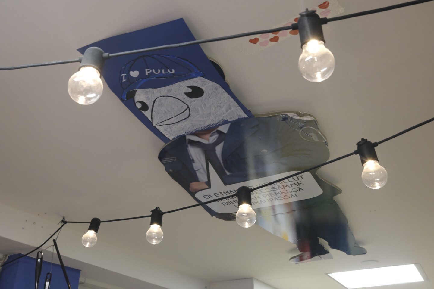 Katonrajasta roikkuva suuri leikattu lintuhahmo, jolla on päänä piirroslintu ja vartalona pukupuku, sininen taustapala jossa näkyy osittainen teksti "I ♥ PULU", ympärillä riippuu mustilla johdoilla sarja paljaita hehkulamppuja.