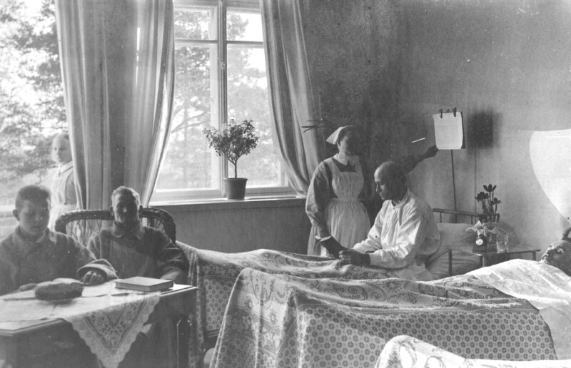 Mustavalkoinen historiallinen valokuva sairaalahuoneesta, jossa potilas makaa vuoteessa, hoitaja seisoo vieressä ja kaksi henkilöä istuu huoneessa, ikkuna taustalla.