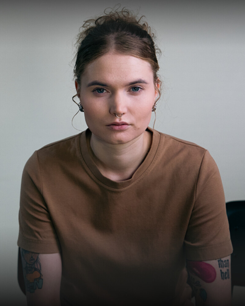 Nuori nainen ruskeassa paidassa, jolla on nenäkoru ja tatuointeja käsivarsissaan, katsoo suoraan kameraan totisella ilmeellä.
