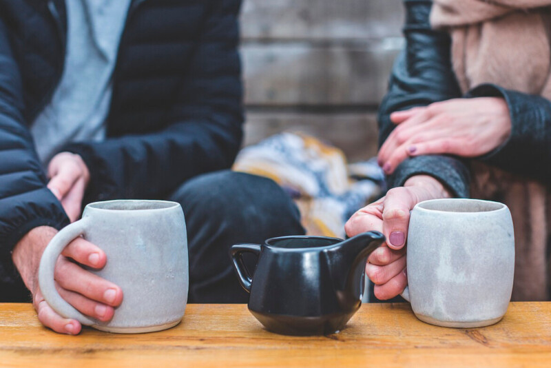 Kuvituskuva: kaksi henkilöä istuu vierekkäin ja pitelee kupeistaan kiinni. Keskellä on teekannu.