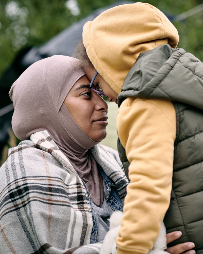 Äiti huivissa ja lapsi koskettavat otsia toisiinsa ulkona, iloinen kohtaaminen.