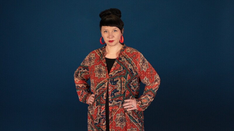 Tummahiuksinen nainen katsoo kameraan kädet lanteilla.