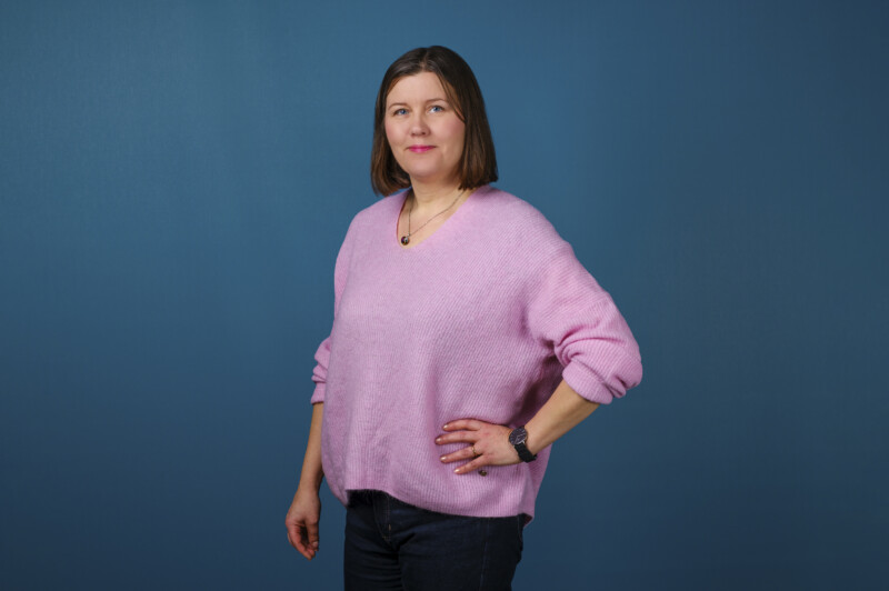 Violettiin neuleeseen pukeutunut nainen katsoo hymyillen kameraan.