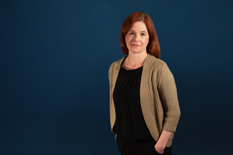 Näkymättömät-ohjelmajohtaja Heidi Rosbäck seisoo sinistä taustaa vasten ja katsoo kameraan