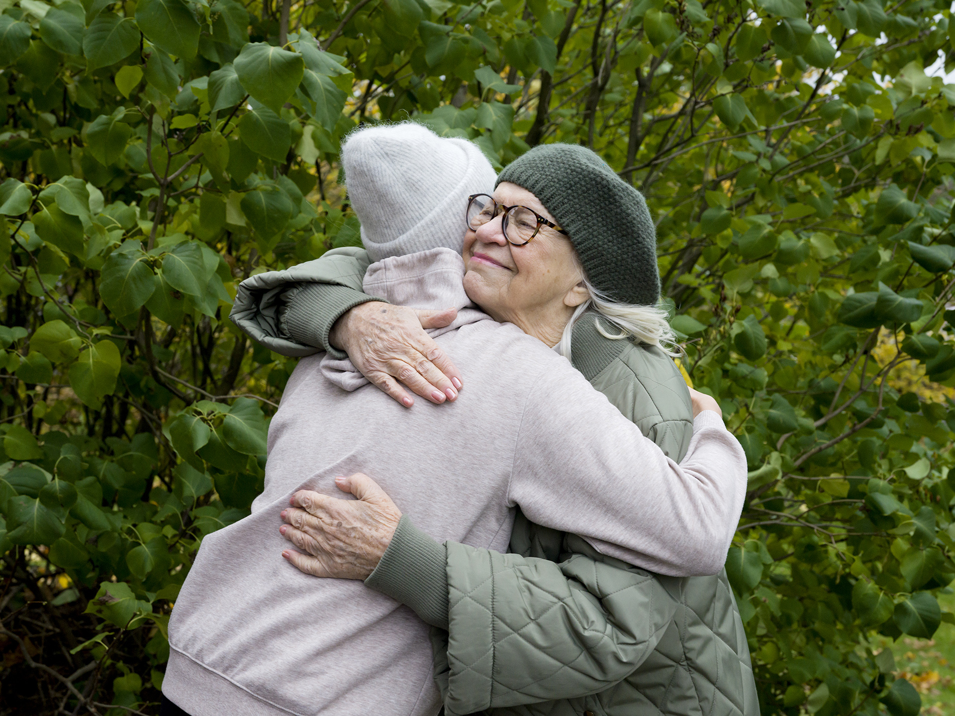 Kaksi naista, toinen iäkkäämpi, halaavat iloisesti toisiaan ulkona vihreiden puiden edessä.