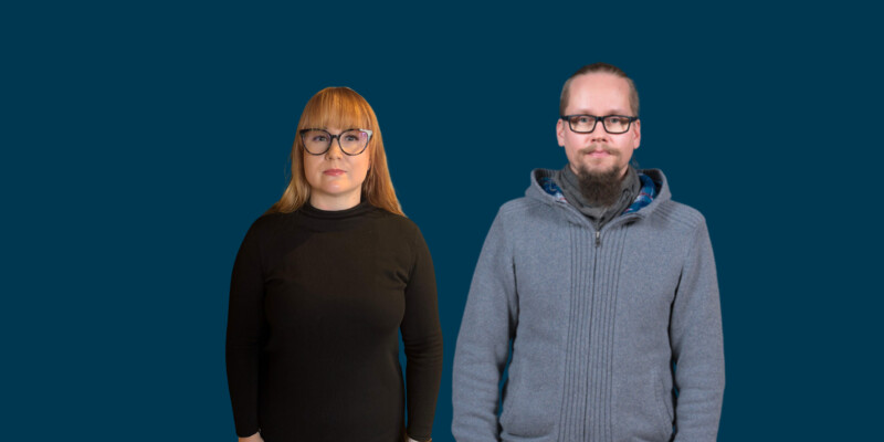 Kaksi henkilöä seisoo sinisen taustan edessä, molemmat katsovat suoraan kameraan; vasemmalla pitkähiuksinen nainen mustassa paidassa ja oikealla lyhyttukkainen mies sinisessä neuleessa ja silmälaseissa.