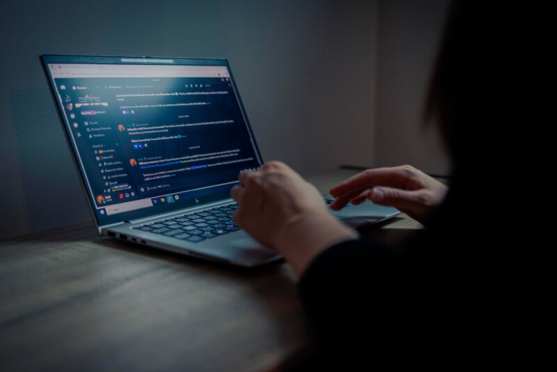 Henkilö on sivuttain kannettavan tietokoneen äärellä siten, että hänen sorrmensa ovat näppäimistön päällä ja ruudulla näkyy keskustelualusta.