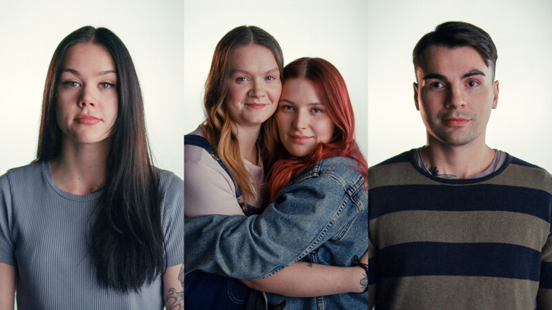 Kolme nuorta aikuista, kaksi naista ja yksi mies, katsovat suoraan kameraan, keskellä olevat naiset hymyilevät ja halaavat toisiaan.