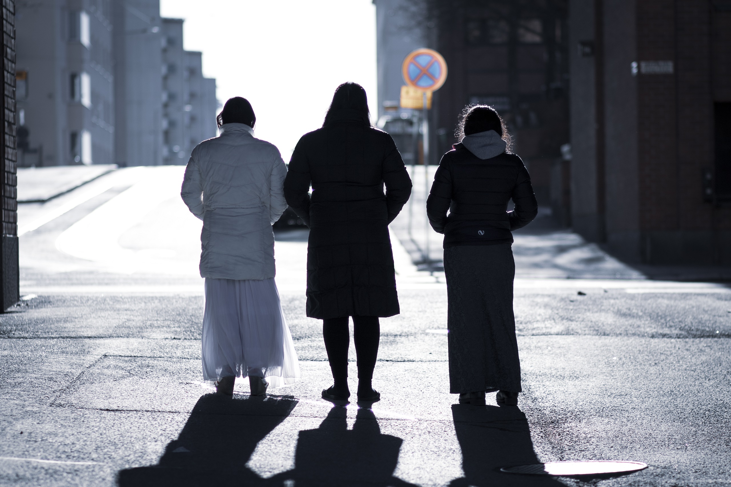 Kolme henkilöä selin kameraan seisomassa auringonvalaisella kadulla, taustalla kaupunkimaisema rakennuksineen.