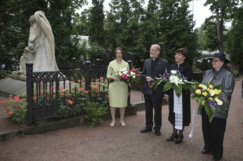 Neljä henkilöä seisoo kukkaseppeleiden kanssa muistomerkillä hautausmaalla, taustalla puita ja pensaita.