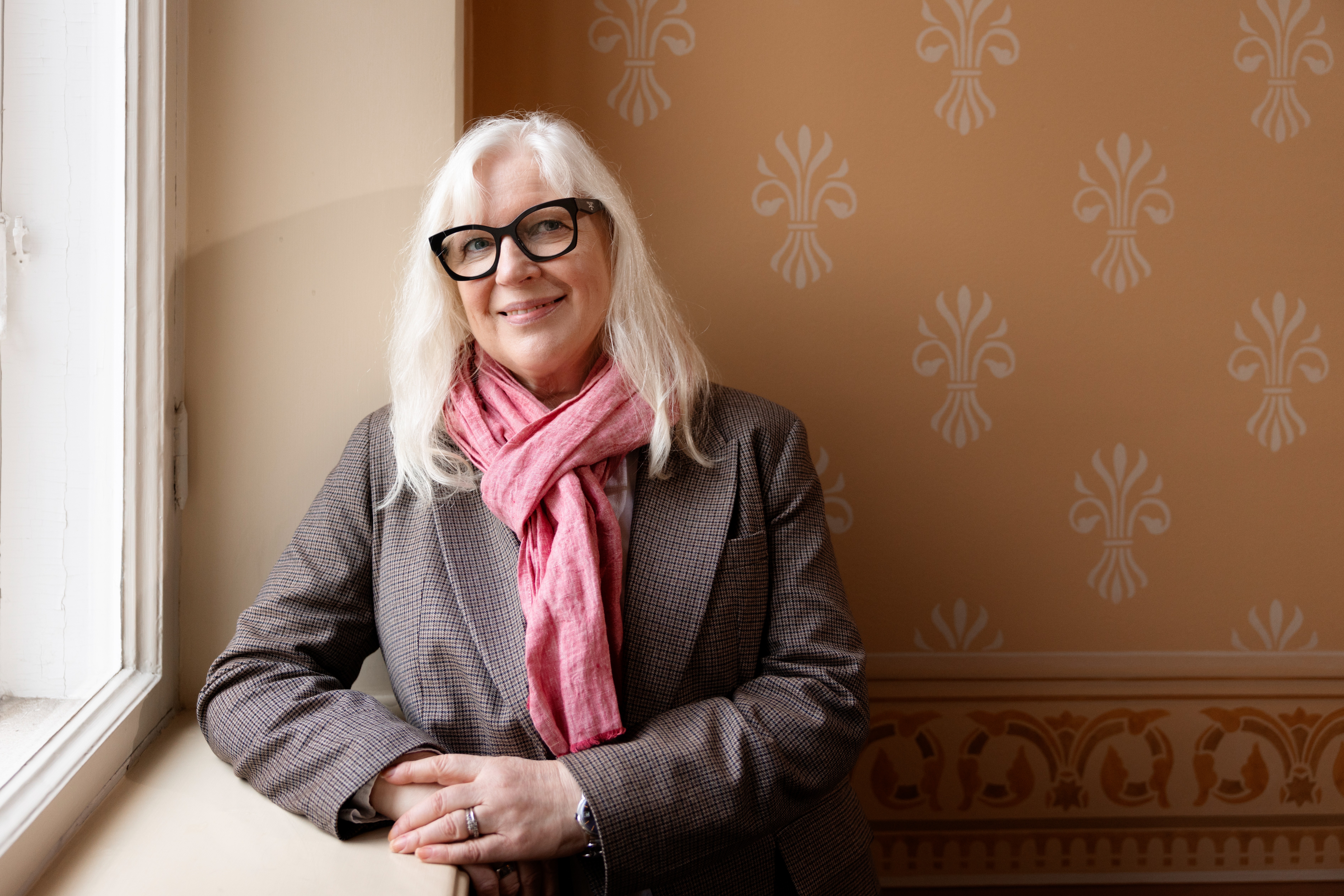 Keski-ikäinen nainen vaaleissa hiuksissa, mustat silmälasit, harmaa takki ja pinkki huivi, nojaa ikkunalaudalle ja hymyilee pehmeässä luonnonvalossa koristeellisen seinän edessä.