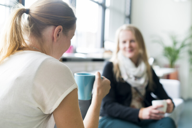 Kaksi naista keskustelee rennosti kahvikupit käsissään valoisassa huoneessa, taustalla ikkunasta tulvii luonnonvaloa.