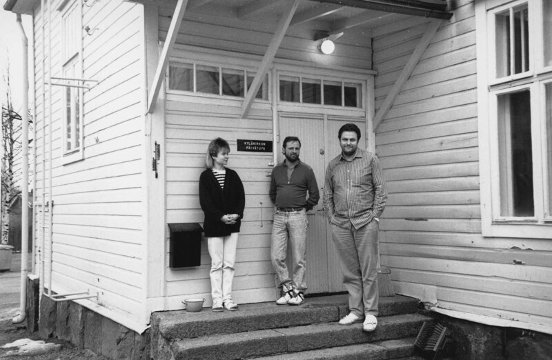 Kolme ihmistä seisoo puisen rakennuksen sisäänkäynnin portailla.