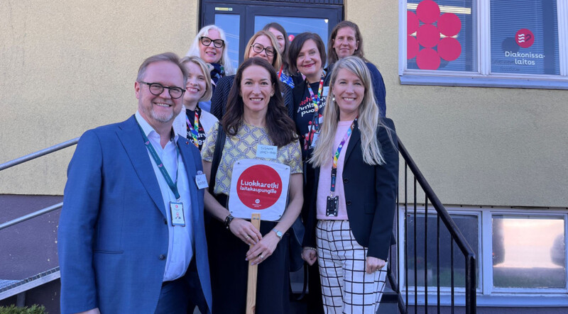 Ryhmä hymyileviä aikuisia seisoo portailla rakennuksen edessä keväisessä ulkoilmassa.