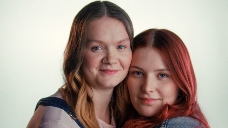 Kaksi nuorta naista nojaamassa lähekkäin ja hymyilemässä lempeästi.