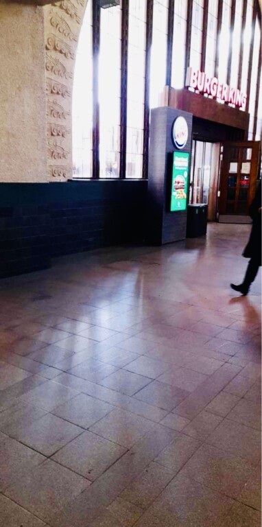 Sisäkuva julkisesta tilasta, jossa näkyy Burger Kingin ravintolan sisäänkäynti ja suuri ikkuna, lattialla heijastuksia ja ihmisen jalat kävelemässä oikealla reunalla.