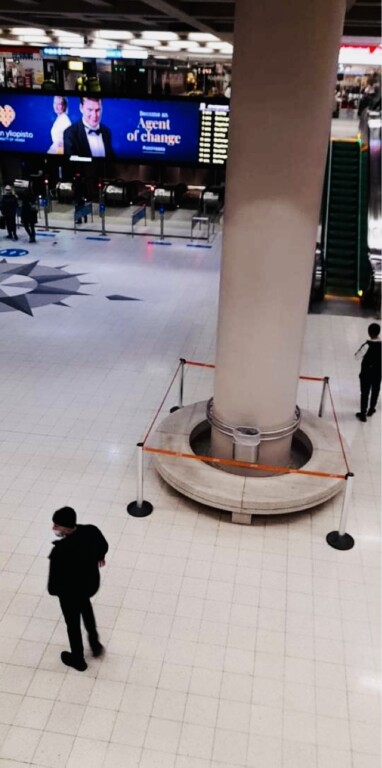 Sisäkuva vilkkaasta julkisesta tilasta, jossa näkyy suuri pylväs, ympärillä esteet, lattialla ihmisiä ja taustalla valotauluja.