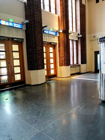 Tyhjä, valoisa sisätila, jossa korkeat ikkunat ja punatiiliset pylväät, sekä kaksi oviaukkoa, joissa on uloskäyntiin viittaavia kylttejä.