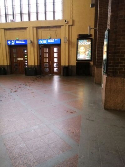 Rautatieaseman sisätila, jossa näkyy kaksi oviaukkoa sinisillä opasteilla ja mainosvalotaulu, lattialla näkyy useita likatahroja ja jälkiä.