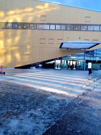 Kuvan etualalla on porrastettu sisäänkäynti moderniin rakennukseen, jonka julkisivu on vaaleaa puuta ja isoja ikkunoita, valoisassa ulkoilmassa.