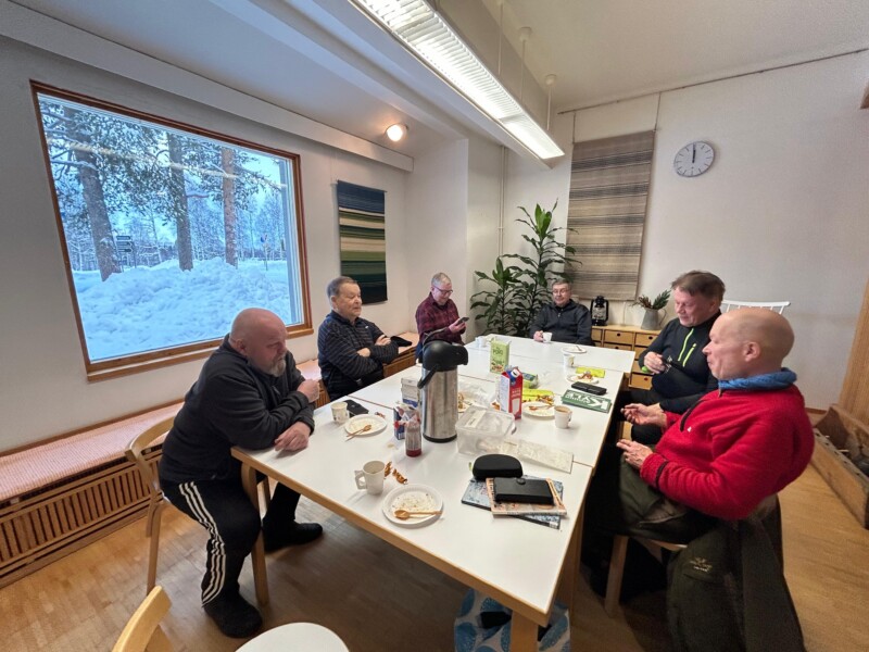 Keskellä huonetta olevan pöydän äärellä istuu kuusi aikuista henkilöä, joista osa katsoo puhelintaan, osa juttelee ja osa kuuntelee. Pöydän keskellä on kahvitermari ja kertakäyttöastioita, huoneen ikkunasta näkyy luminen metsämaisema.