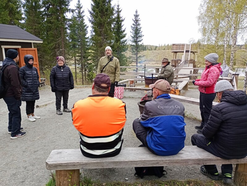 Ihmisiä istuu ja seisoo ympyrässä luontopysähdyspaikalla lintutornin läheisyydessä. Lintutornin opastaja seisoo ringin keskellä ja puhuu.