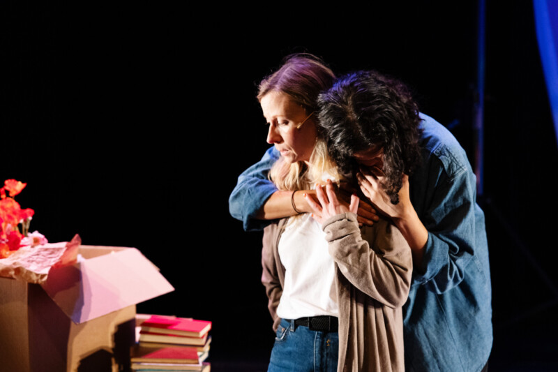 Kaksi näyttelijää halaavat toisiaan koskettavassa kohtauksessa näyttämöllä, taustalla punaisia kukkia ja pino kirjoja.