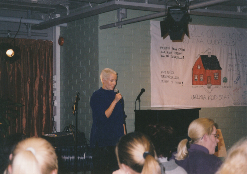 Sisäkuva, jossa henkilö puhuu mikrofoniin yleisölle, taustalla seinällä juliste, jossa piirros talosta ja tekstiä.