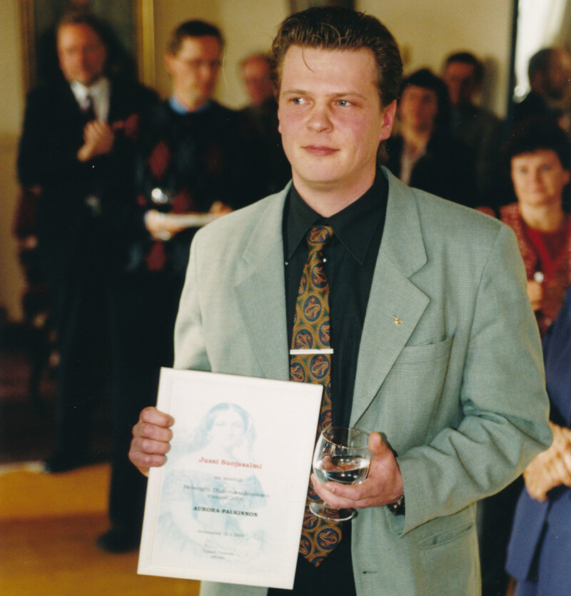 Mies seisoo juhlatilaisuudessa kädessään kunniakirja ja lasi, taustalla muita ihmisiä.