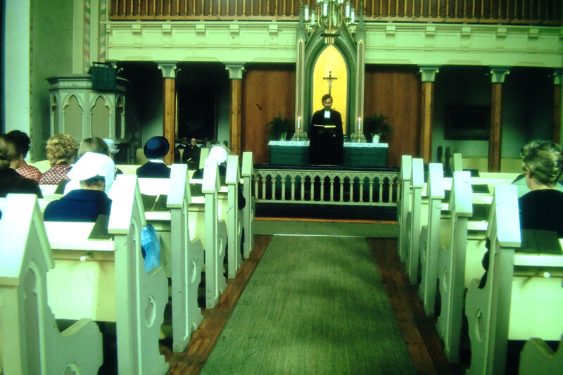 Kirkon sisäkuva 1970-luvulta, jossa papin johtama jumalanpalvelus ja seurakuntalaisia penkeissä.