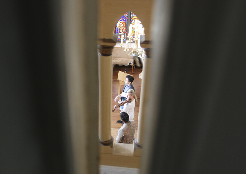 Kirkon sisäkuva, jossa näkyy kolme henkilöä, joista yksi soittaa viulua, ja taustalla värikkäitä lasimaalauksia.