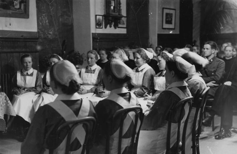 Mustavalkoinen historiallinen valokuva, jossa useita diakonissoja ja miehiä istumassa piirissä huoneessa, kaikki pukeutuneina aikakauden asuihin.