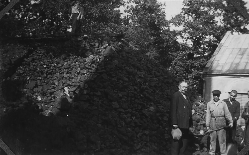 Mustavalkoinen valokuva, jossa neljä miestä seisoo kiviaidan vieressä ulkona, taustalla puita ja rakennus.