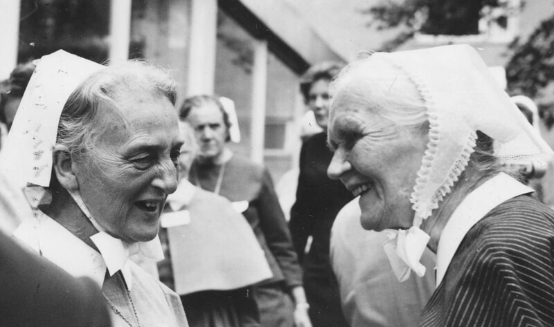 Mustavalkoinen valokuva kahdesta hymyilevästä vanhemmasta naisesta, jotka ovat pukeutuneet diakonissojen asuihin, taustalla ihmisiä ja rakennus.