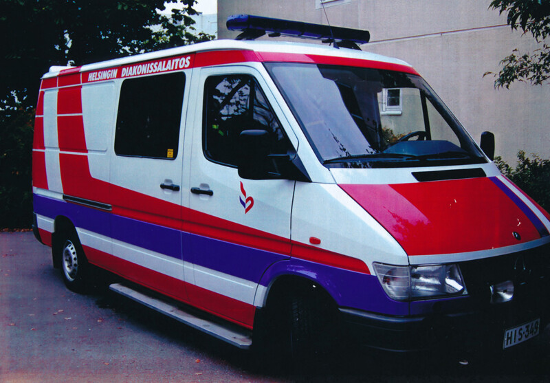 Liikkuva ambulanssi, jossa on punavalkoinen Diakonissalaitoksen tunnus ja sininen hälytysvalo katolla, pysäköitynä rakennuksen edessä.