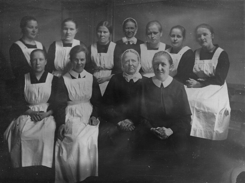Mustavalkoinen ryhmäkuva, jossa kymmenen naista istuu ja seisoo rivissä, kaikki pukeutuneina vanhanaikaisiin diakonissan asuihin ja esiliinoihin.